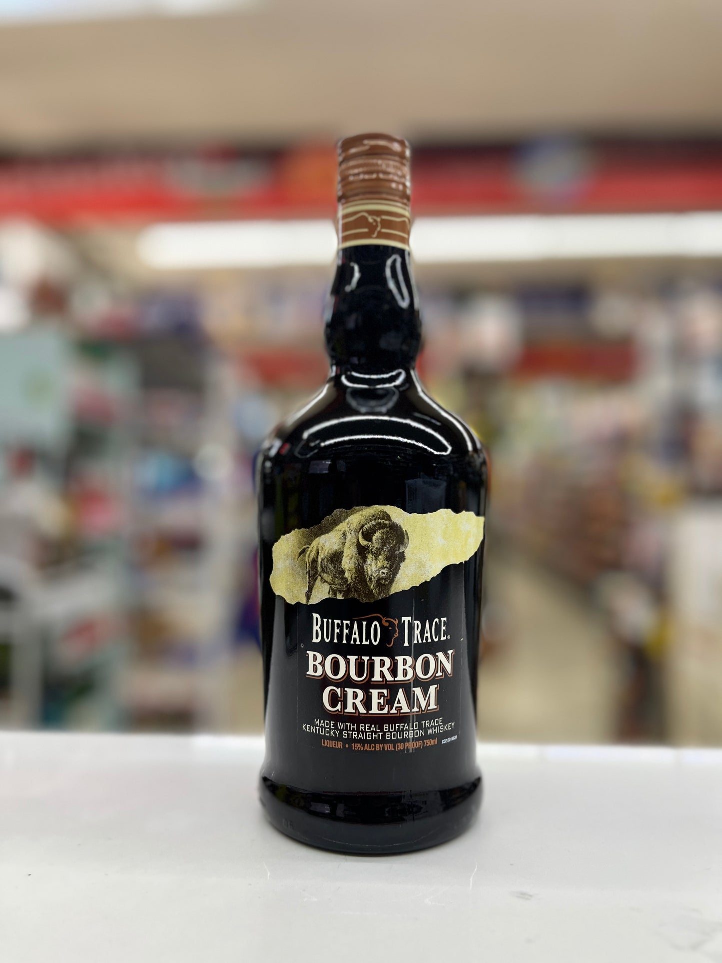 A bottle of Buffalo Trace Bourbon Cream whiskey stands on a white surface with a blurred store background. The bottle is dark glass with a gold label and a brown wax seal at the neck.