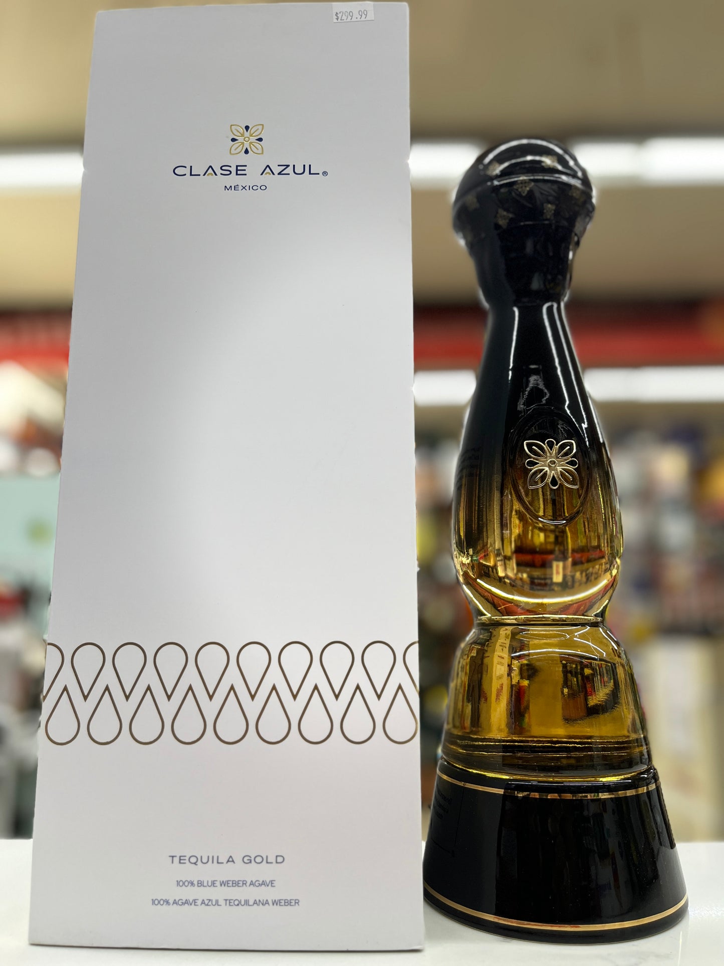 A bottle of Clase Azul Tequila Gold next to its tall white packaging box, set against a blurred retail background.
