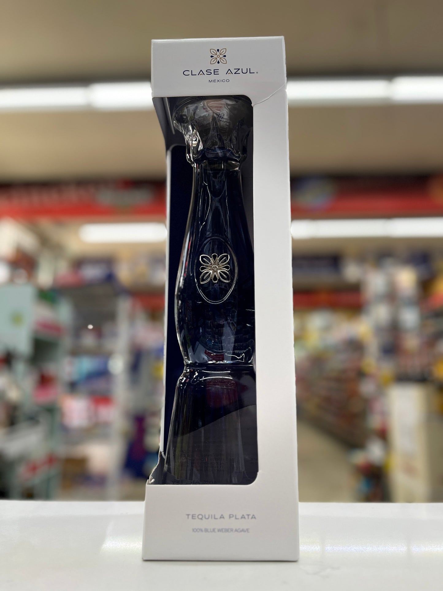 A boxed bottle of Clase Azul Tequila Plata, shown in its white retail packaging with a clear window displaying the dark blue bottle, set against a blurred store background.