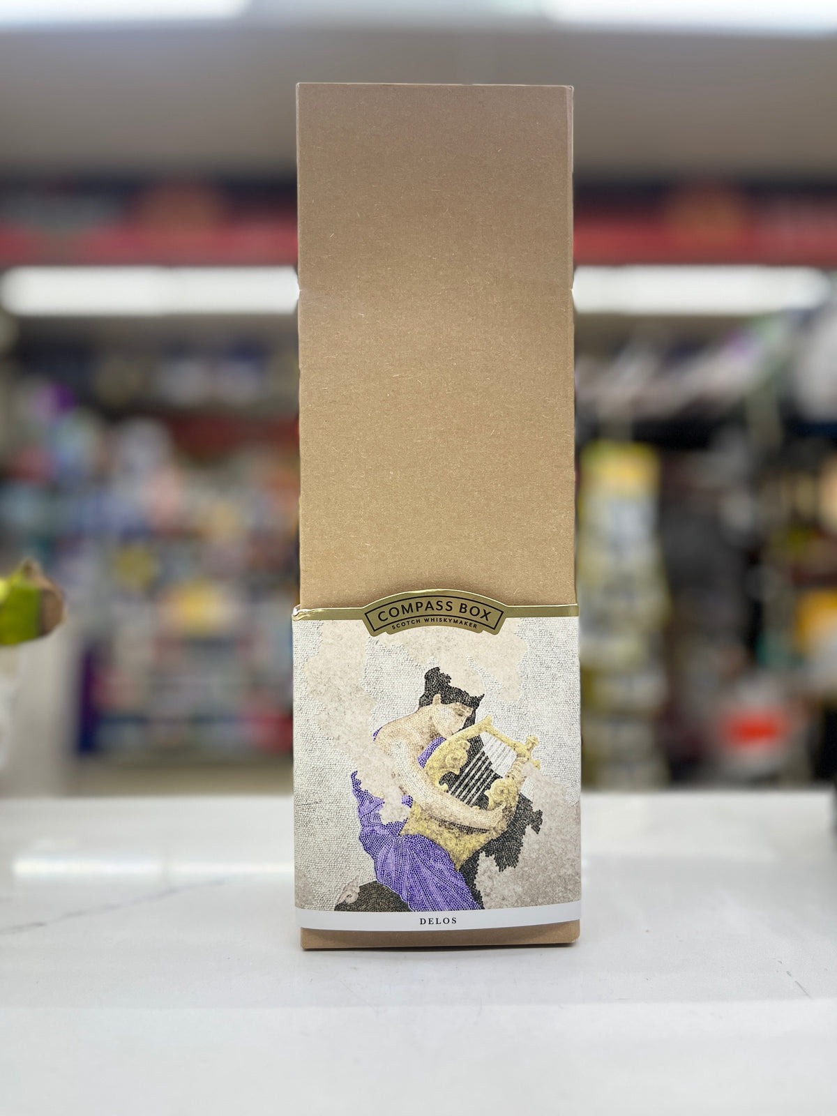 A tall, narrow brown cardboard box with a decorative label near the bottom, standing upright on a white surface with a blurred store background. The label shows an illustration of a figure playing a lyre and includes the text 'COMPASS BOX DELOS'.