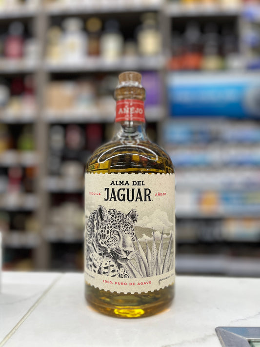 A bottle of Alma Del Jaguar Anejo tequila standing on a white counter with a blurred liquor store shelf background.