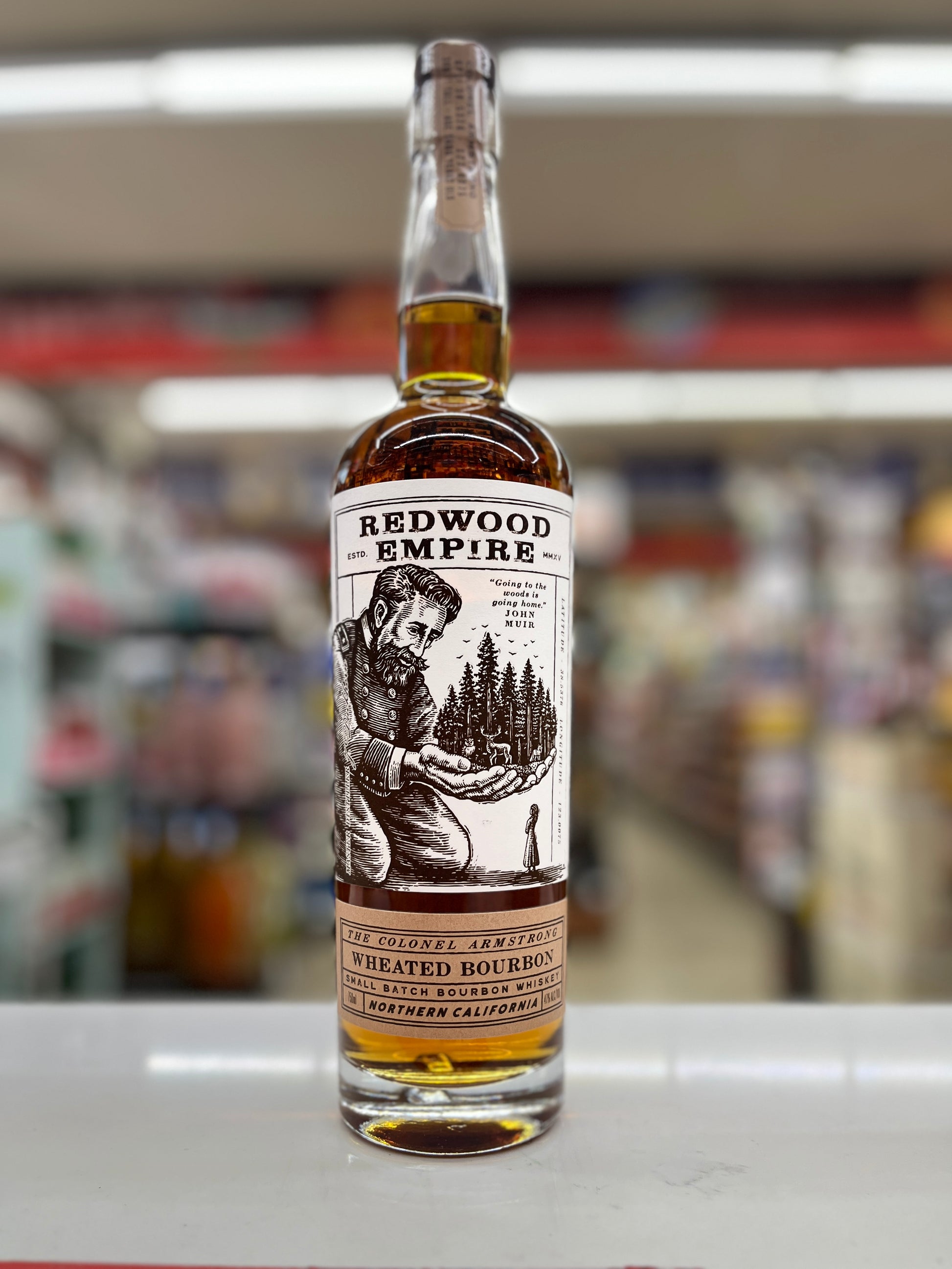 A bottle of Redwood Empire wheated Bourbon Whiskey called The colonel armstrong on a white shelf, with a blurred store background.