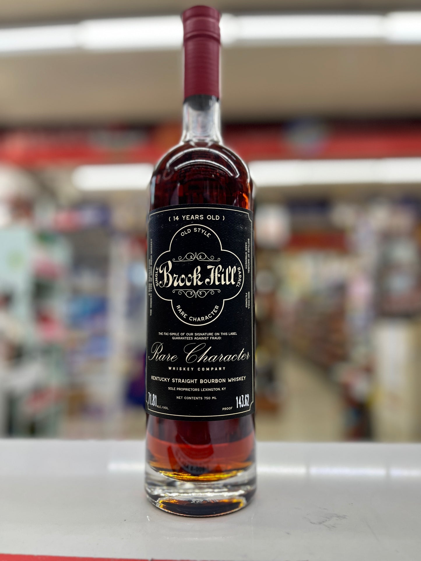 A bottle of Brook Hill 14 Years Kentucky Straight Bourbon by Rare Character Whiskey. This bottle is called pulled together standing on a white counter in a store, with a blurred background of store shelves.