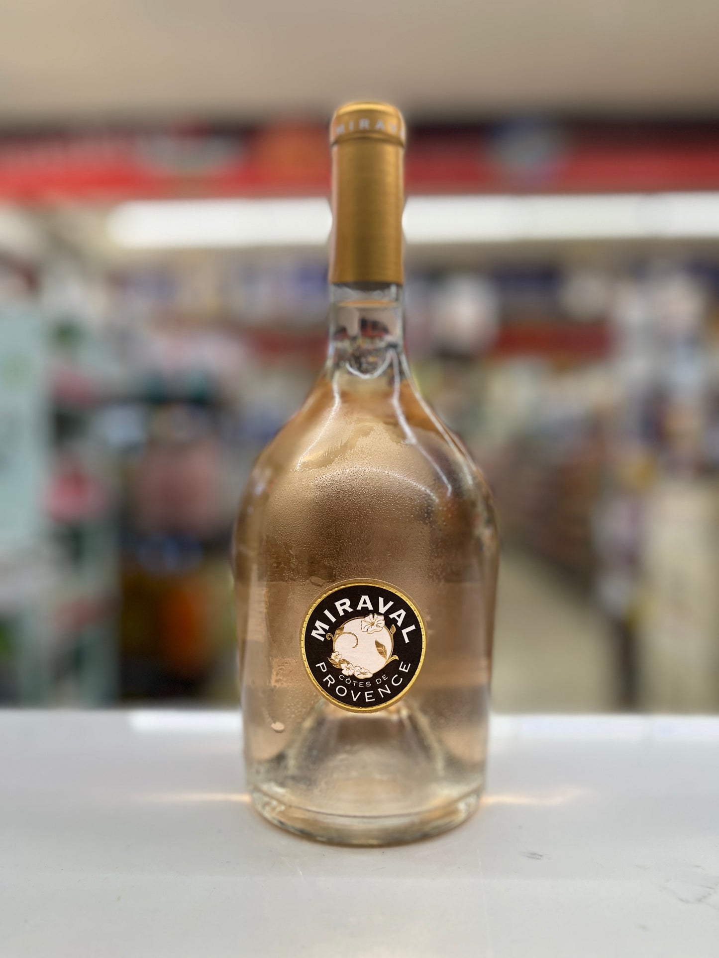 A bottle of Miraval Provence rosé wine with a gold foil cap and a circular black, white, and gold label, sitting on a white surface with a blurred store background.