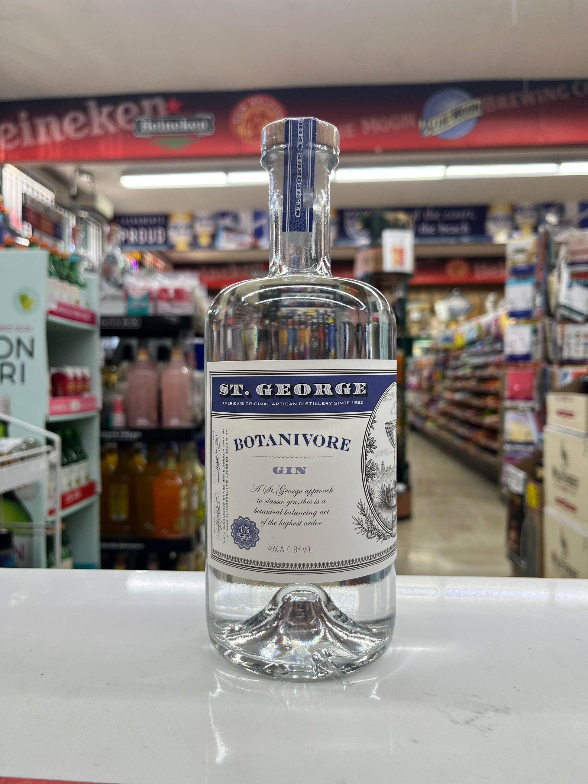 A bottle of St. George Botanivore Gin on a white counter inside a retail store, with shelves and aisles blurred in the background.