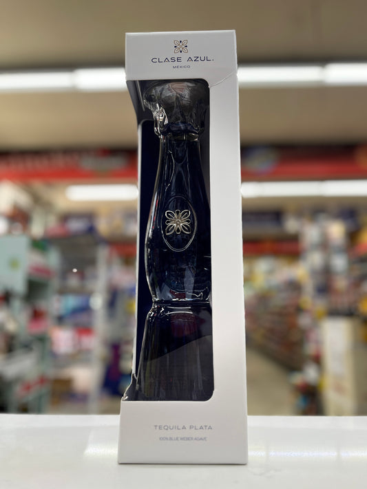 A boxed bottle of Clase Azul Tequila Plata, shown in its white retail packaging with a clear window displaying the dark blue bottle, set against a blurred store background.