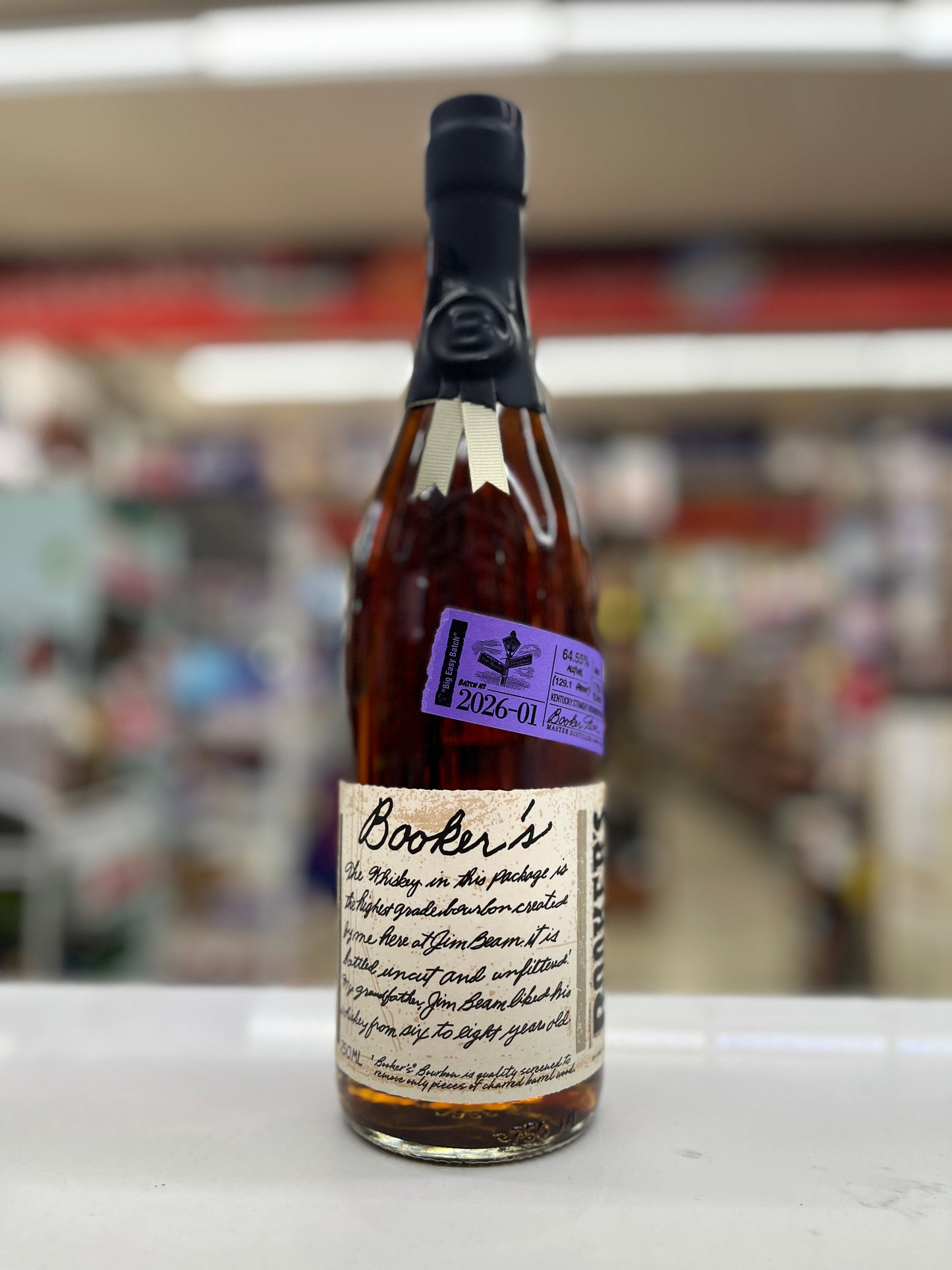 A bottle of Booker's Bourbon "Big Easy Batch" whisky on a white surface with a blurred shop background. The bottle has a black neck seal, amber liquid, a cream label with black script, and a purple sticker.
