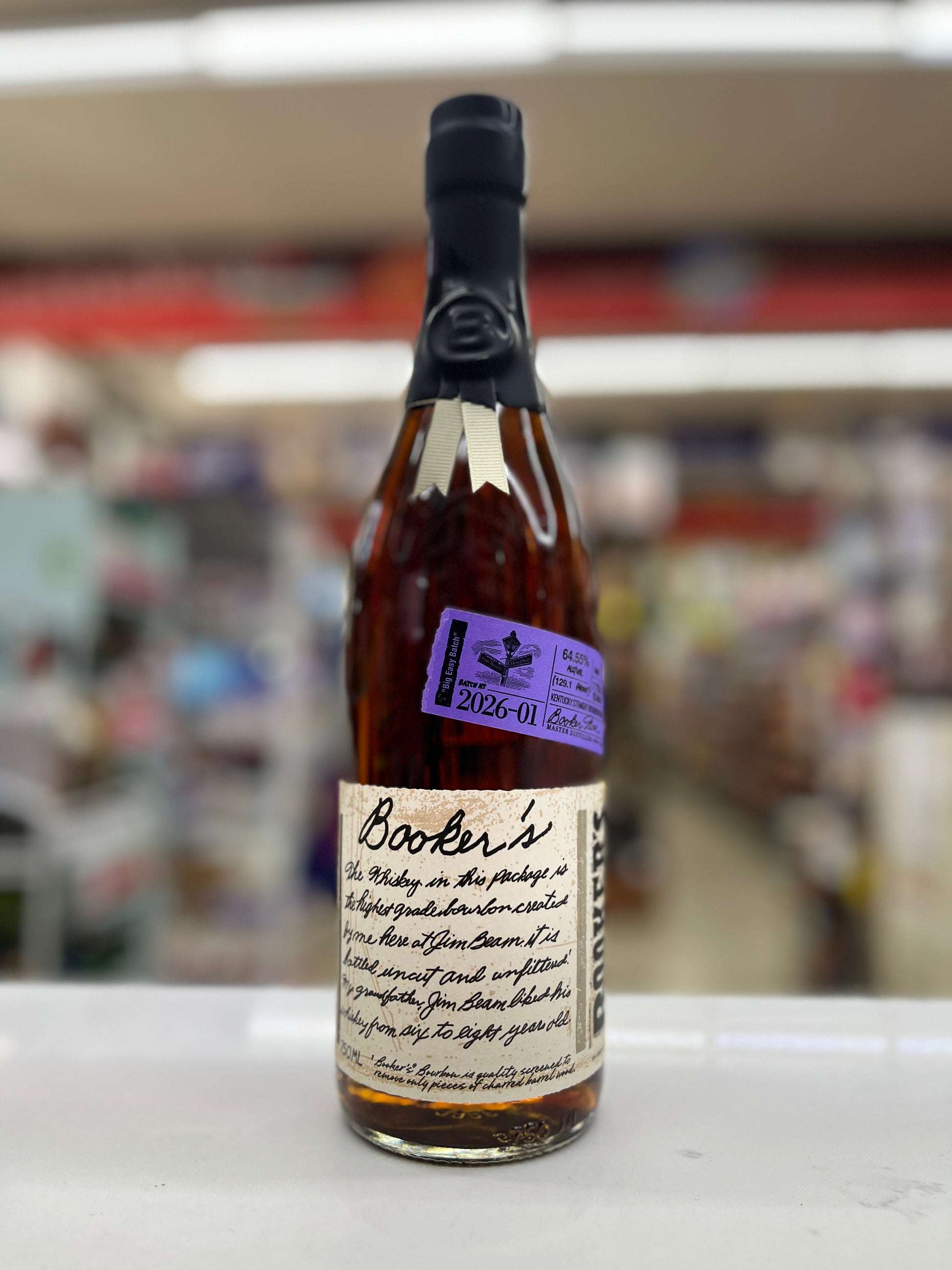 A bottle of Booker's Bourbon "Big Easy Batch" whisky on a white surface with a blurred shop background. The bottle has a black neck seal, amber liquid, a cream label with black script, and a purple sticker.