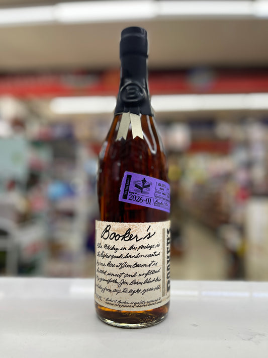 A bottle of Booker's Bourbon "Big Easy Batch" whisky on a white surface with a blurred shop background. The bottle has a black neck seal, amber liquid, a cream label with black script, and a purple sticker.