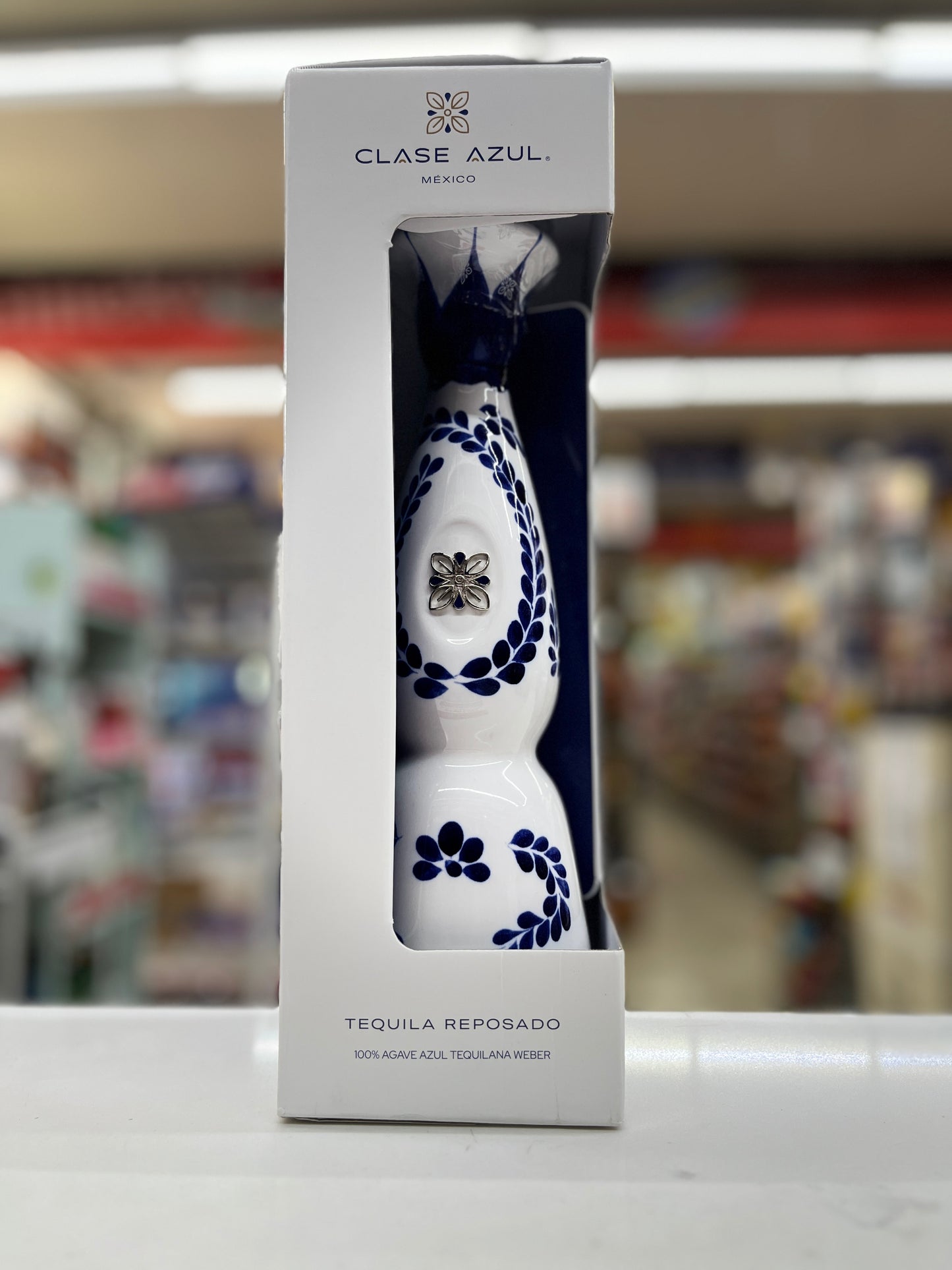 A boxed tequila bottle from Clase Azul Reposado 375ml on a countertop with a blurred store background. The product is a white tequila bottle with dark blue floral and vine patterns, presented inside a tall white retail box.
