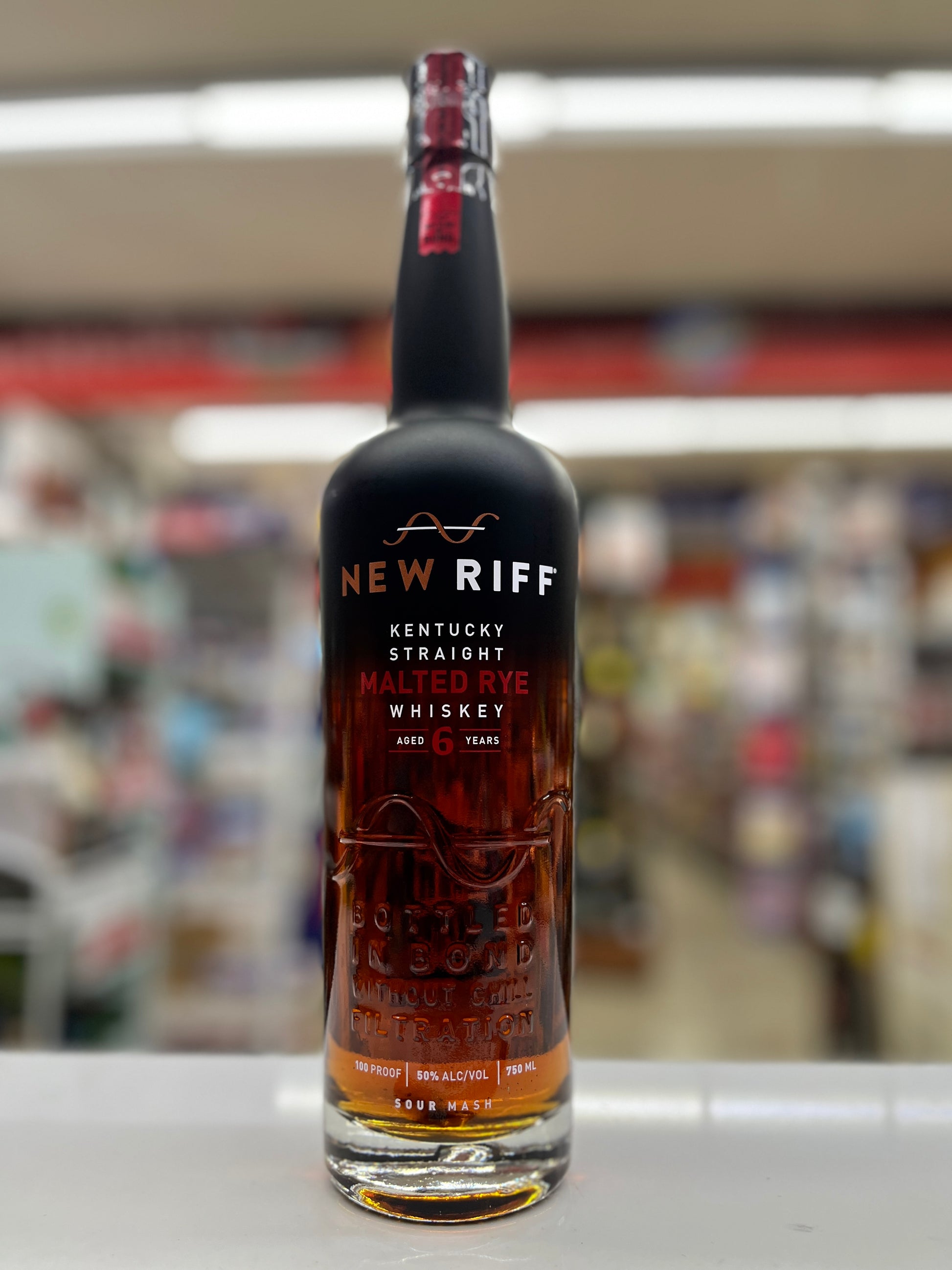A bottle of New Riff Kentucky Straight Malted Rye Aged 6 years Whiskey standing on a white counter in a blurred retail store environment.
