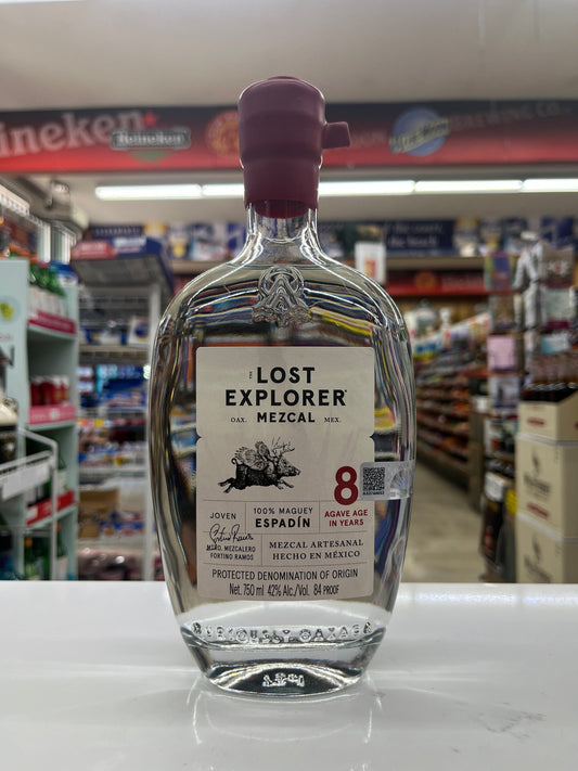 A bottle of Lost Explorer Espadin Mezcal made by using 8 years old agave plant displayed on a white counter inside a retail store with blurred shelves and signage in the background.