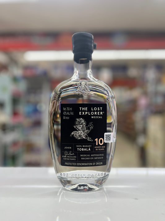 A bottle of The Lost Explorer Mezcal Toblal 10 Year Old Agave used featuring a clear glass bottle with a black label and black cap, set against a blurred store background.