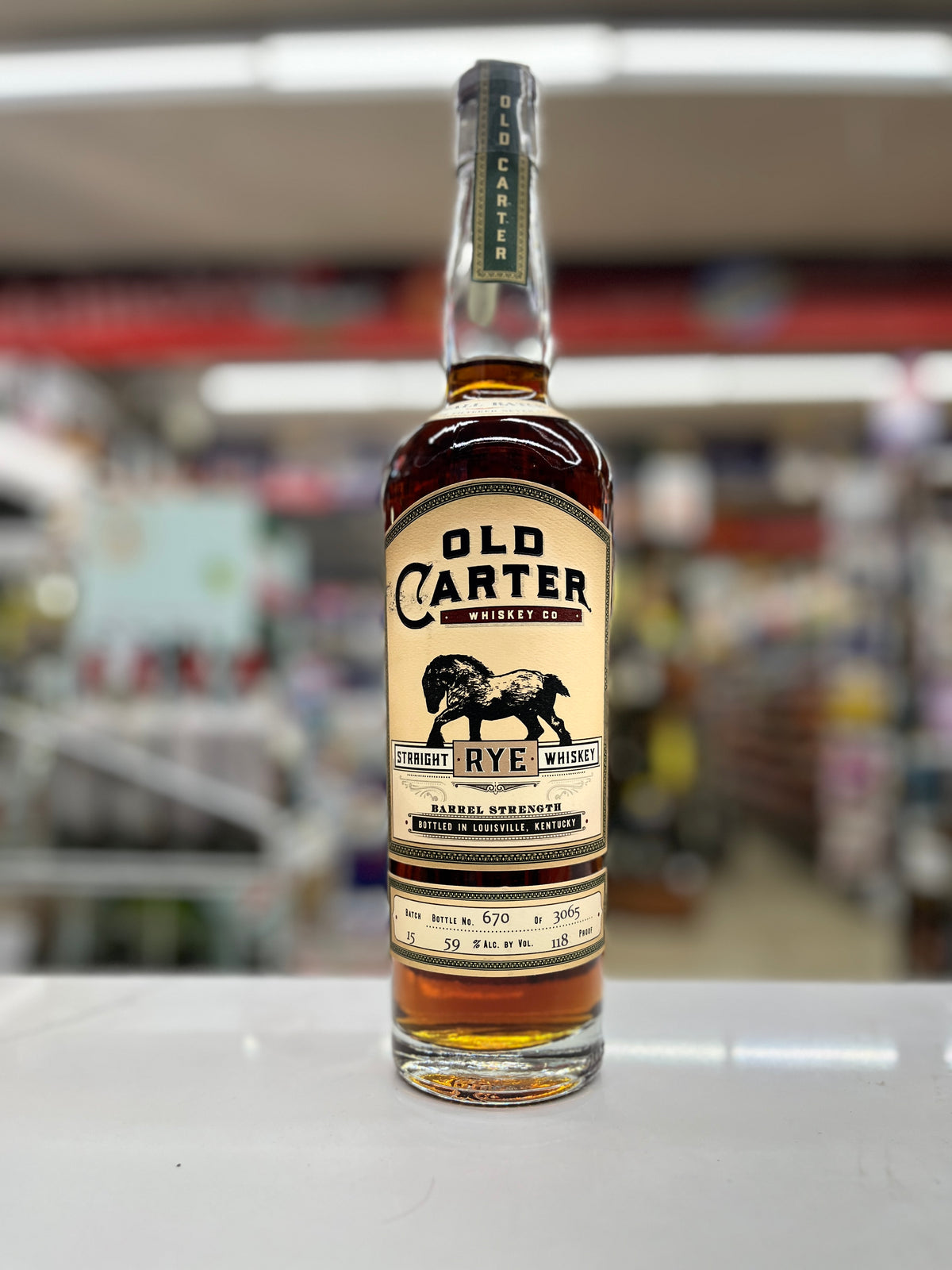 A bottle of Old Carter Rye Whiskey standing on a white counter in a blurred retail store environment. The bottle has a cream-colored label with black text and a black buffalo graphic, and the amber liquid inside is visible through the clear glass.