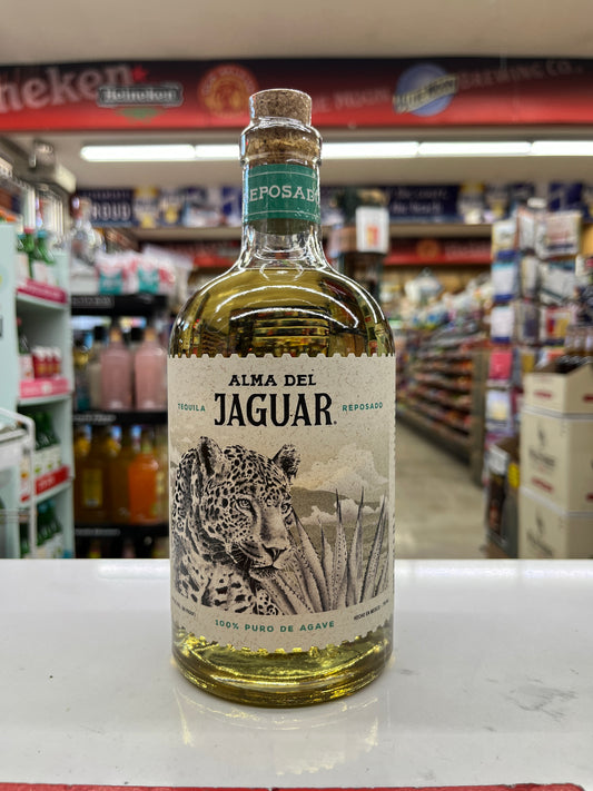 A bottle of Alma Del Jaguar Reposado tequila standing on a white counter inside a retail store. The clear glass bottle shows the golden-yellow aged tequila inside, with a cork stopper and a cream-colored label featuring a black and white jaguar illustration.
